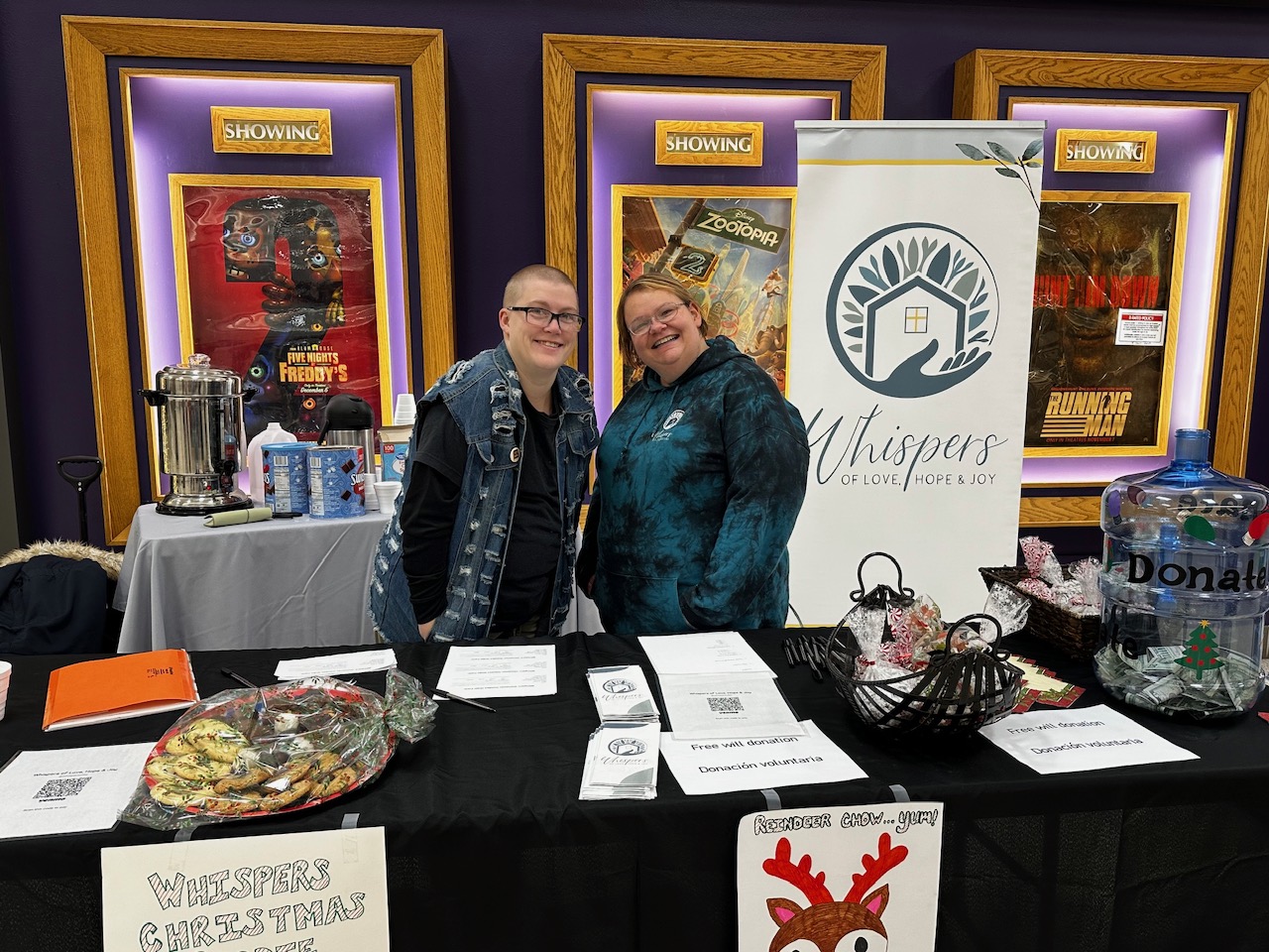 Two women behind a Whispers table with cookies and a donation jar in a theater lobby