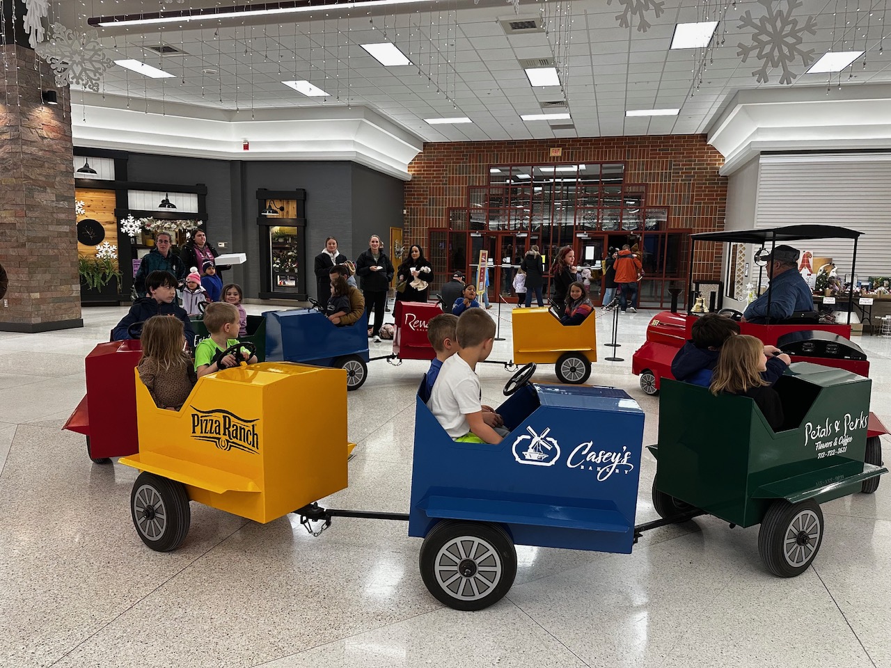 Children riding colorful cars inside a shopping center, with adults standing nearby