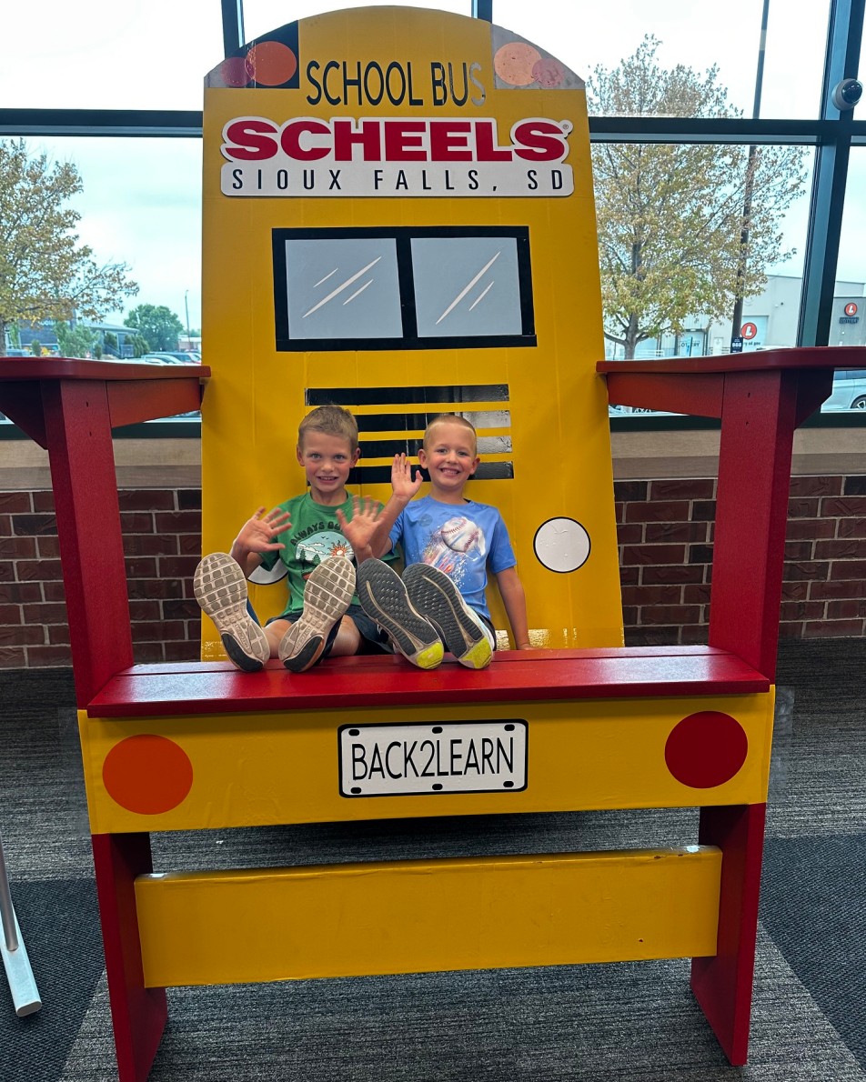 Two boys waving from an oversized yellow school-bus chair labeled Back2Learn