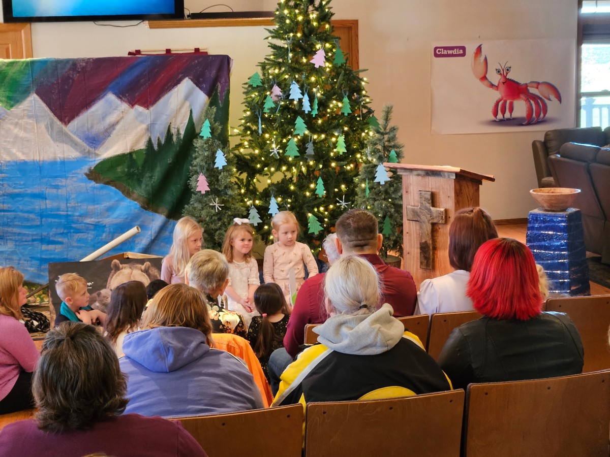 Adults watch children standing in front of a lit Christmas tree and a painted backdrop