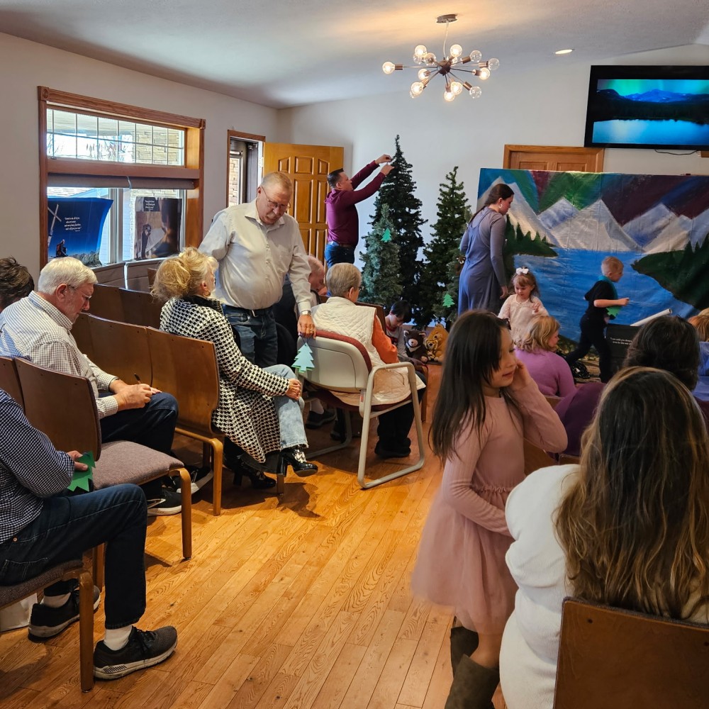 Adults and children in a worship room while people decorate small Christmas trees