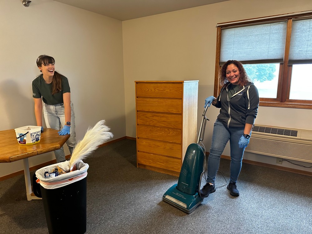 Two people cleaning a carpeted room, one with a vacuum and one beside a table