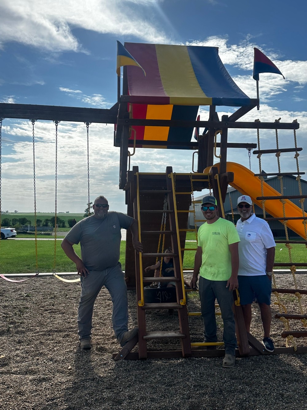 Three adults pose in front of a wooden playground while a child sits on the structure