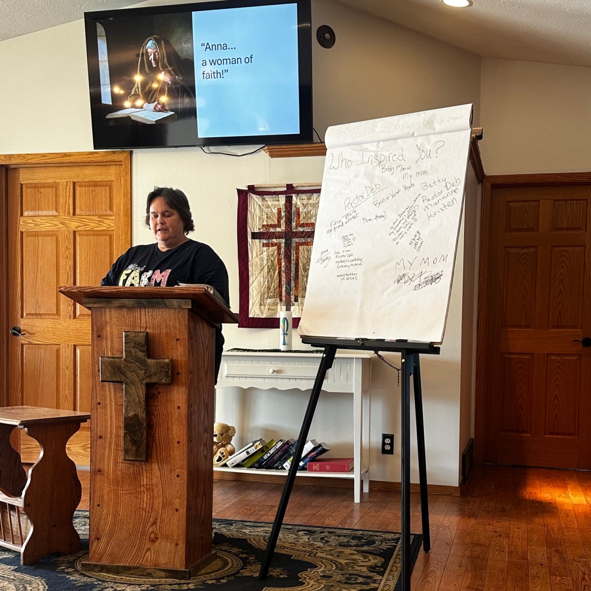 A woman at a wooden pulpit beside a flip chart, with a lesson on the screen behind her