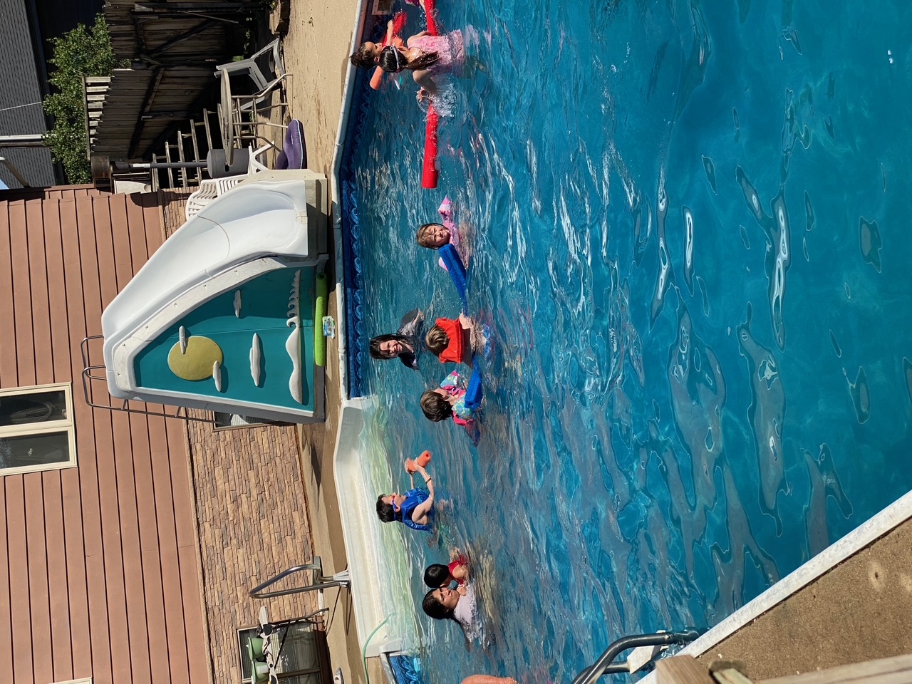 Adults and children in a backyard pool, some in life jackets, with a slide behind them