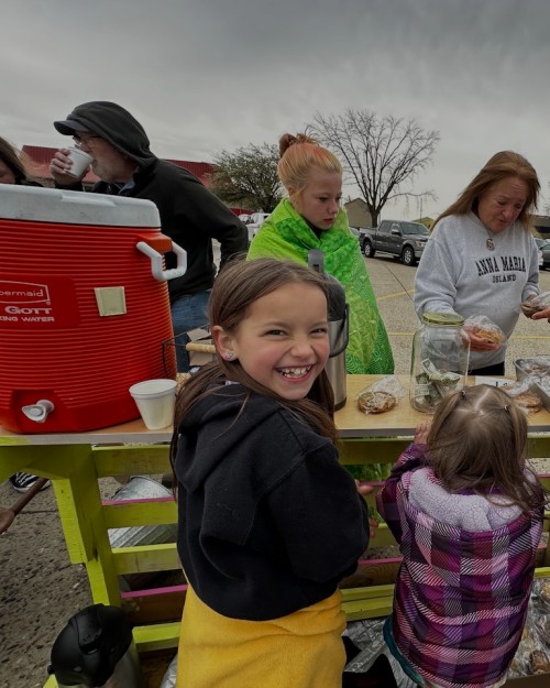 A child smiling at an outdoor table with a drink cooler, snacks, and adults behind the table