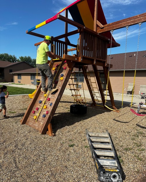 A person in a yellow shirt climbing a wooden playground, with a ladder on the ground and a child nearby